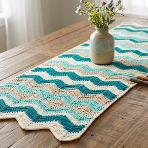 Puede incluir: Camino de mesa tejido a mano con ganchillo, con un patrón de chevron en tonos turquesa, beige y crema. Se muestra sobre una mesa de madera rústica, con un jarrón de flores y ganchillos. También hay un ovillo de lana.