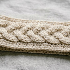 Op de afbeelding: Een beige, handgebreide haarband met een kabelpatroon. De haarband is gemaakt van dik, getextureerd garen. Dit winteraccessoire is ontworpen om de oren warm te houden. Het kabelpatroon voegt een vleugje elegantie toe.