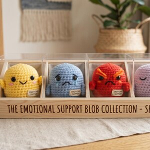 Op de afbeelding: De Emotional Support Blob Collection - Serie 1. Vier gehaakte blobs in een heldere acryl behuizing. Elke blob vertegenwoordigt een emotie: geel (blij), blauw (verdrietig), rood (boos) en paars (slaperig).