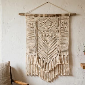 Op de afbeelding: Een crèmekleurige macramé wandhanger met geometrische patronen en franjes. De textielkunst is opgehangen aan een houten deuvel, wat een vleugje bohemien toevoegt aan een ruimte met neutrale tinten. Een ingelijste botanische print en een potplant zijn ook zichtbaar.