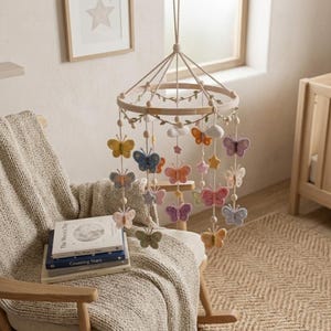 Peut inclure: Mobile pour bébé en bois avec des papillons, des étoiles et des nuages en feutre colorés. Suspendu au-dessus d'un fauteuil à bascule avec une couverture et des livres. Une figurine de renard en bois est sur une étagère. Ambiance chambre d'enfant.