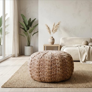 May include: A cozy living room scene featuring a large, round, brown knitted pouf on a jute rug. A beige sofa, a wooden side table with a vase of dried flowers, and a potted plant are also visible. The room has a neutral color palette.