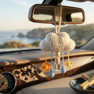 Puede incluir: Un adorno de coche con forma de nube de ganchillo, de color crema, cuelga del espejo retrovisor de un coche. El adorno tiene detalles de ganchillo amarillos y azules, incluyendo flores, estrellas y gotas. Una pequeña botella de cristal está suspendida debajo.