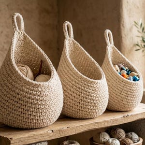 Puede incluir: Tres cestas de ganchillo beige en forma de lágrima con asas de bucle están sobre una estantería de madera. Las cestas están llenas de hilo, materiales de manualidades y juguetes pequeños. Un cuenco de hilo está debajo.
