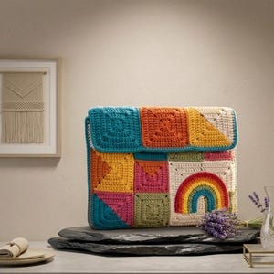 May include: A colorful, handmade crochet bag with a rainbow design. The bag features a patchwork design with squares of teal, orange, yellow, pink, and white. The bag is displayed on a dark gray stone.