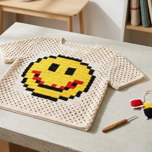 May include: Cream-colored, crocheted t-shirt featuring a pixelated smiley face design in yellow, red, and black. The shirt is displayed on a light gray surface, with crochet supplies including yarn, thread, and a crochet hook.