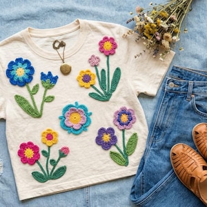 Puede incluir: Camiseta color crema adornada con flores de ganchillo coloridas, combinada con vaqueros azules, sandalias marrones y un bolso tote tejido. Un collar con un colgante está en la camisa. También hay flores secas.