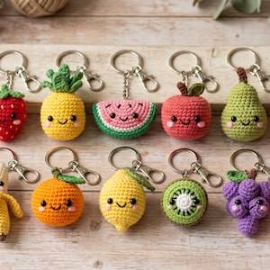Könnte beinhalten: Eine Sammlung von gehäkelten Obst-Schlüsselanhängern, darunter eine Erdbeere, Ananas, Wassermelone, Apfel, Birne, Kirschen, Banane, Orange, Zitrone, Kiwi, Trauben und Pfirsich. Jeder Anhänger hat ein lächelndes Gesicht und einen silbernen Schlüsselring.