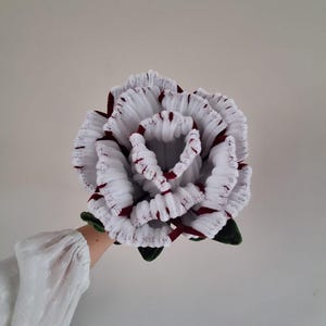 May include: A handcrafted rose flower made from white and dark red yarn. The petals feature a textured, ribbed appearance with white edges and deep red centers. Dark green leaves are attached at the base of the flower head.