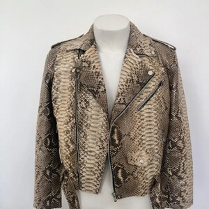 Puede incluir: Una chaqueta estilo motero con un estampado de piel de serpiente en tonos beige y marrón. La chaqueta incluye un cuello con solapas, cremalleras metálicas plateadas en la parte delantera y bolsillos, un bolsillo pequeño con solapa y botón.