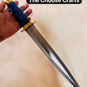 Può includere: Un coltello a lama fissa con una lunga lama in acciaio appuntita, una guardia in ottone e un manico cilindrico con venature del legno blu e nere. Il pomolo è in ottone e il testo The Choose Crafts è visibile nell'angolo in alto a destra.