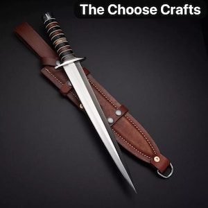 Può includere: Un pugnale a doppio taglio con lama in argento lucido e impugnatura con anelli alternati neri e marroni. Il pugnale poggia su un fodero in pelle marrone con cuciture e rivetti metallici visibili. Il testo The Choose Crafts è visibile.