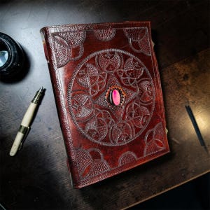 May include: A dark brown leather journal with an embossed Celtic knot design and a pink gemstone. The journal is closed and lies on a wooden surface, next to a pen and inkwell. The cover features intricate patterns.