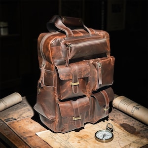 May include: A brown leather backpack with multiple pockets and a top handle. The bag has a rugged, vintage look with visible stitching and metal hardware. A compass and rolled map sit on a wooden surface in the foreground.