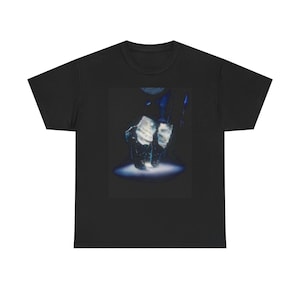 May include: Black t-shirt featuring a graphic of a person's feet in black shoes, illuminated by a spotlight. The image has a dark, artistic aesthetic. The t-shirt is made of a soft material.