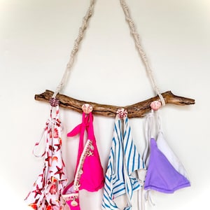 May include: A driftwood wall hanging displaying four bikini tops. The tops are in various colours and patterns, including a white and red starfish print, hot pink, blue and white stripes, and lavender. The driftwood is suspended by a macrame rope.