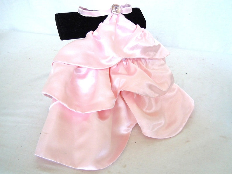 May include: A pink satin ruffle collar with a rhinestone embellishment. The collar is displayed on a black velvet stand.