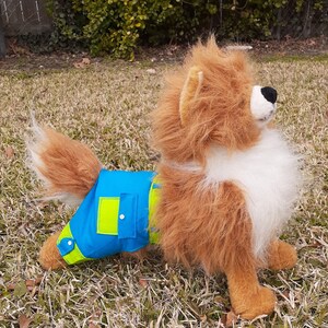 May include: A plush toy dog wearing a blue and green dog diaper. The diaper has a pocket on the side.