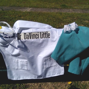 May include: A child's doctor costume set, including a white lab coat with pockets and the text "Dr. DaVinci Little", teal scrubs, and a yellow and clear medical device. The costume is laid out on a dark surface with green grass in the background.
