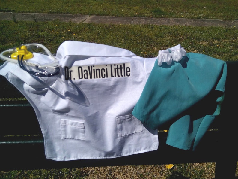 May include: A child's doctor costume set, including a white lab coat with pockets and the text "Dr. DaVinci Little", teal scrubs, and a yellow and clear medical device. The costume is laid out on a dark surface with green grass in the background.