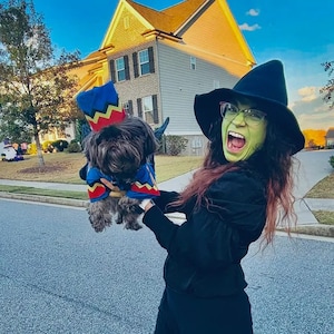 May include: A person with green face paint and a black witch hat wearing a black long-sleeved top, holding a small dog dressed in a blue, red, and yellow costume with a matching hat. The dog has dark fur and is wearing a colorful patterned outfit.
