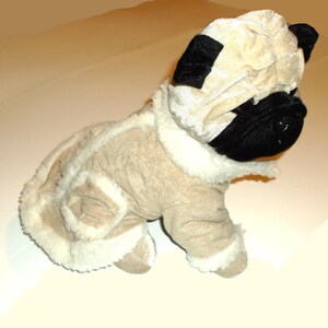 Puede incluir: Un juguete de peluche de un carlino que lleva un abrigo de piel marr&oacute;n y blanco con capucha. El juguete tiene ojos negros y una nariz negra.