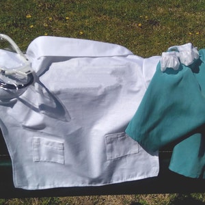 May include: A white medical lab coat with two pockets and a teal scrub set. A stethoscope with a yellow and clear plastic head is draped over the coat. The items are arranged on a dark green bench.