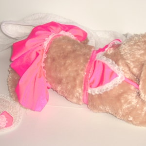 May include: A plush dog wearing a pink and white bikini and a pink skirt. The dog is lying on its side with its head resting on its paw. The dog is wearing a white crocheted hat.