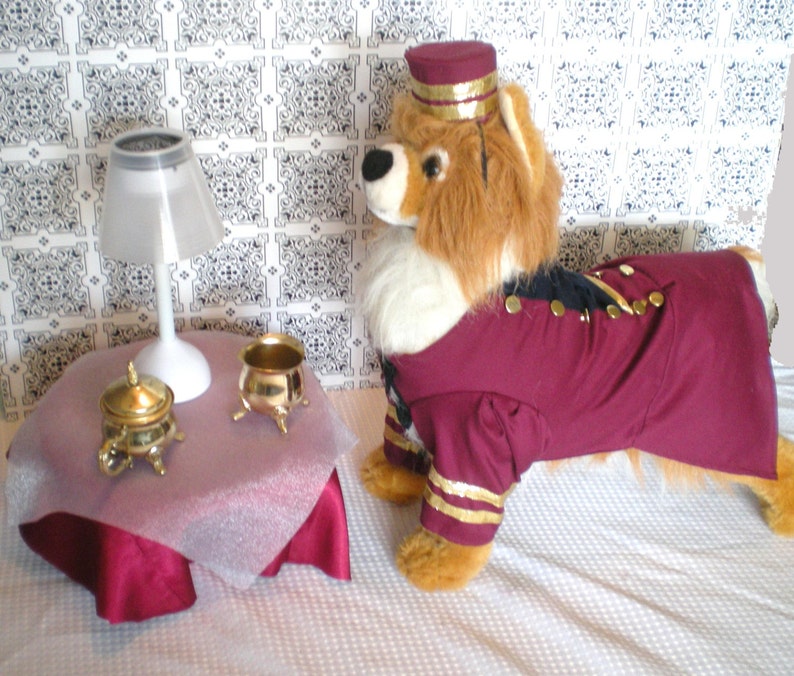 May include: A plush lion wearing a burgundy and gold uniform with a matching hat. A small table with a white lamp and gold-colored decorative items is in the foreground. The background features a patterned wall.