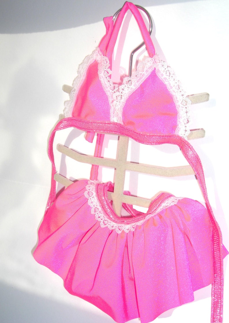 May include: Pink sparkly bikini top and skirt with white lace trim. The top has a halter style and the skirt is a mini skirt.
