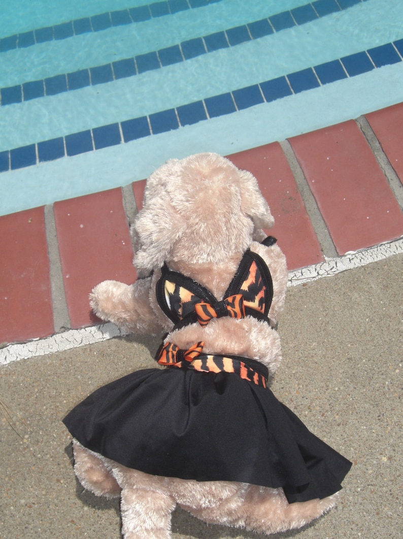 May include: A plush toy dog wearing a black skirt and a butterfly-patterned swimsuit. The dog is sitting on a concrete surface near a swimming pool.