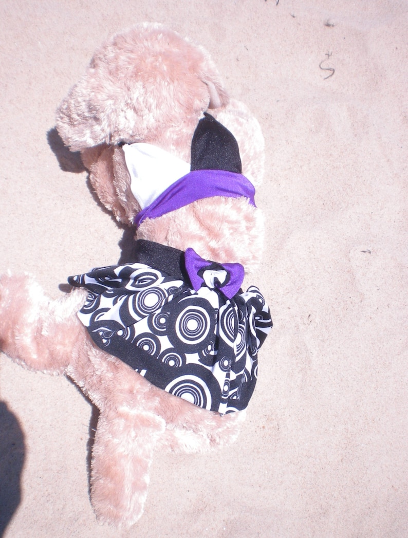 May include: A plush toy wearing a purple and black bikini top and a black and white skirt with a circle pattern. The toy is lying on its back on a sandy surface.