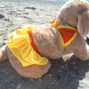 May include: A plush dog wearing a yellow and orange swimsuit. The dog is lying on a sandy beach.