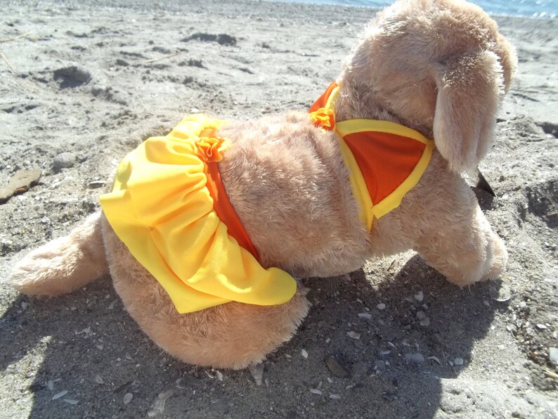 May include: A plush dog wearing a yellow and orange swimsuit. The dog is lying on a sandy beach.