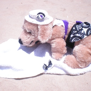 May include: A plush dog wearing a white sun hat with a purple flower, a purple and white striped halter top, and a black and white patterned skirt. The dog is lying on a white towel.