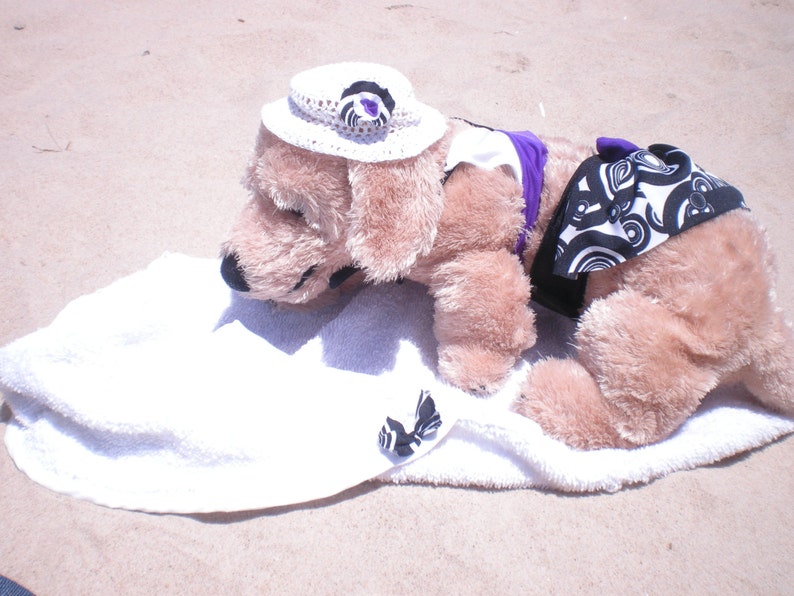 May include: A plush dog wearing a white sun hat with a purple flower, a purple and white striped halter top, and a black and white patterned skirt. The dog is lying on a white towel.