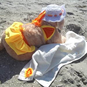 May include: A small, light brown dog wearing a yellow and orange swimsuit and a white crocheted sun hat. The dog is lying on a white towel on a sandy beach.
