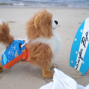 May include: A plush dog with brown and white fur wearing blue swim trunks with a shark pattern and orange side panels, standing next to a blue and white surfboard with the text Surfer Pup and small white flower graphics printed on it.