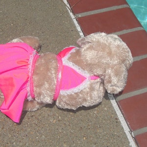 May include: A plush toy dog wearing a pink and white swimsuit. The dog is lying on a concrete surface near a swimming pool.