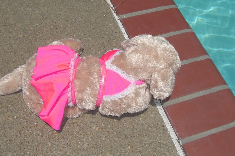 May include: A plush toy dog wearing a pink and white swimsuit. The dog is lying on a concrete surface near a swimming pool.