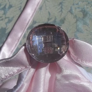 May include: A close-up of a large, clear, faceted, purple glass button. The button is sitting on a pink satin fabric.