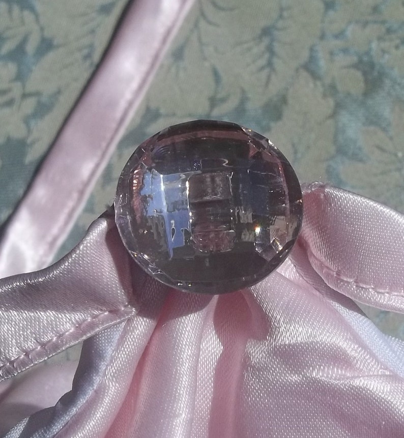 May include: A close-up of a large, clear, faceted, purple glass button. The button is sitting on a pink satin fabric.