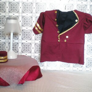 May include: A burgundy-colored costume jacket with gold accents, including buttons and sleeve stripes, hangs against a patterned backdrop. A small table with a lamp and a matching burgundy hat with gold trim are also visible, suggesting a themed display.