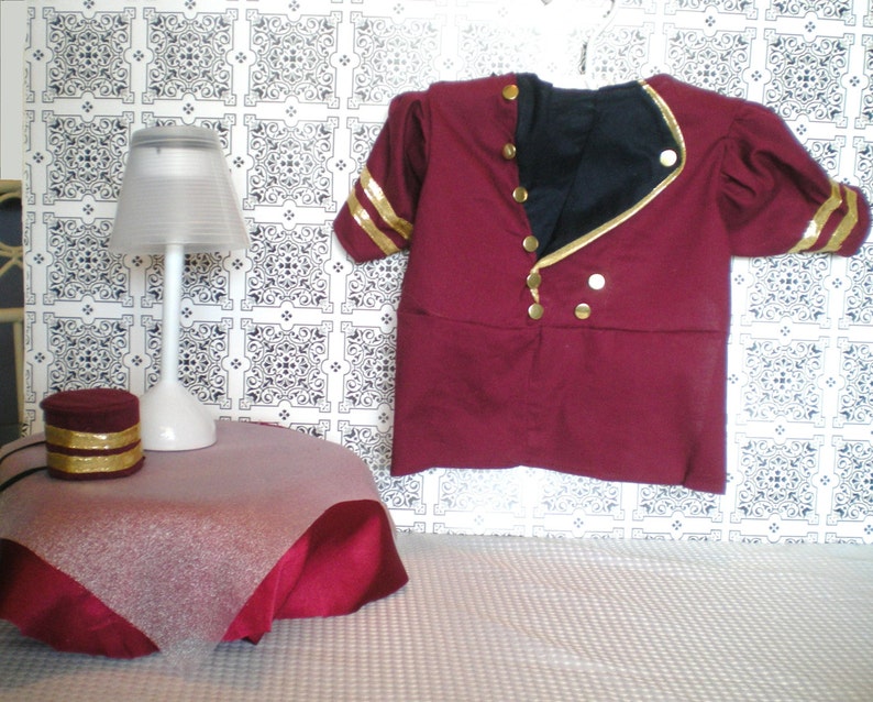 May include: A burgundy-colored costume jacket with gold accents, including buttons and sleeve stripes, hangs against a patterned backdrop. A small table with a lamp and a matching burgundy hat with gold trim are also visible, suggesting a themed display.