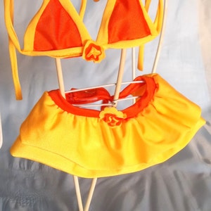 May include: A yellow and orange bikini with a ruffled skirt. The bikini top is a triangle shape and the skirt has a flower embellishment.