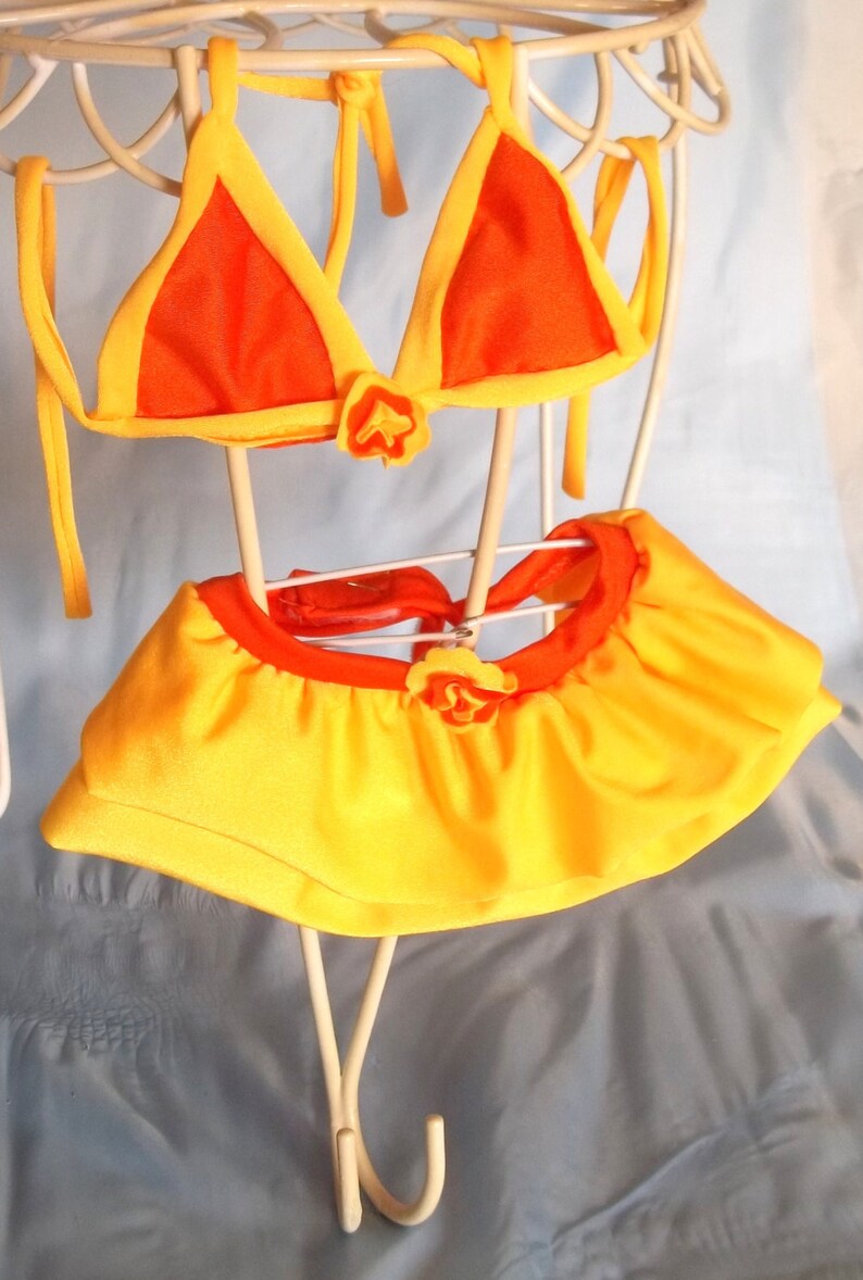 May include: A yellow and orange bikini with a ruffled skirt. The bikini top is a triangle shape and the skirt has a flower embellishment.