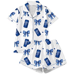 May include: A white pajama set consisting of a short-sleeved button-down shirt and shorts. The fabric features a repeating pattern of blue bows, small blue hearts, and blue beverage cans with the text "I love you" printed on each can.