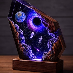 May include: A rectangular resin and wood lamp featuring a space scene with two white astronaut figures, a blue and green Earth, a purple swirling black hole, and a small shooting star, all illuminated from within by a light source in the base.