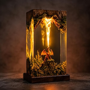 May include: A rectangular resin lamp featuring a wooden base and top, containing a miniature orange dragon perched on a rocky cliff. The dragon breathes a stream of bright yellow light upward toward a cavernous ceiling, with a castle visible nearby.