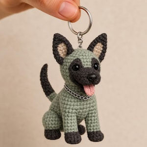 May include: A crochet keychain featuring a small dog with sage green fur, dark charcoal ears, paws, and muzzle. The dog has black bead eyes, a pink tongue, and a silver-toned metal chain collar. It is attached to a circular silver-toned metal keyring.